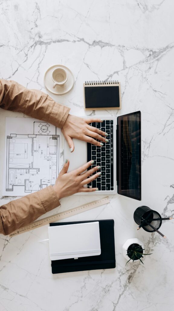 Overhead view of architect using laptop with blueprints and coffee on marble desk.